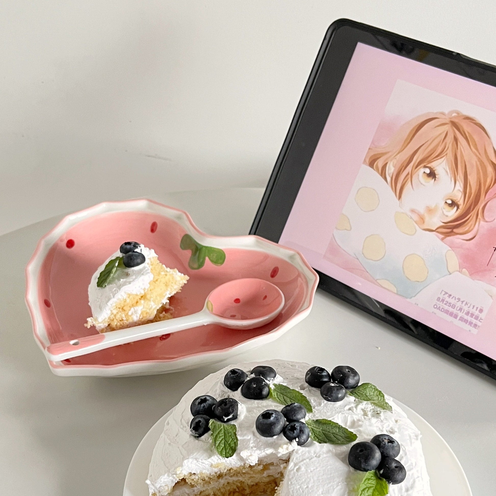 Pie with whipped cream and blueberries on a white plate, with a pink dish and tablet in the background.
